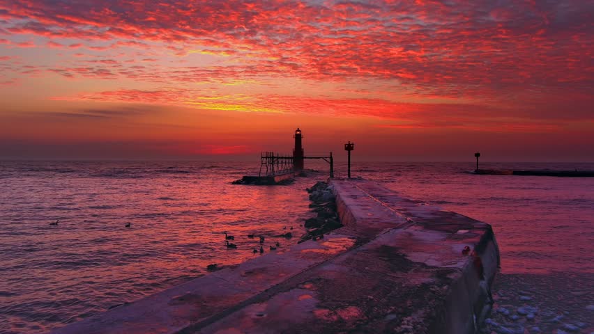 Iconic Lighthouse beacon silhouetted against fiery Lake Michigan twilight horizon, moving aerial view.