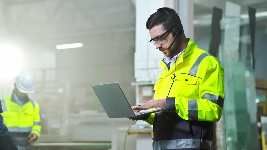 Caucasian male technician in headset and uniform standing in factory, talking with video and working at laptop. Producing concept. Indoor. Man working and videochatting on computer at production. Royalty-Free Stock Footage #1099117425