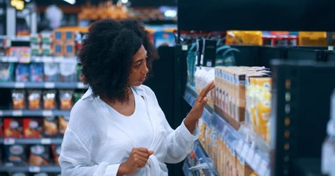 African American young woman shopper, in white casual shirt and blue denim jeans, attentively reading the content on label while shopping for cereals in the grocery store, videoclip de stoc