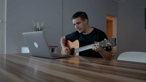 Man plays guitar watching educational videos on laptop. Beginner musician learns to use musical instrument sitting at wooden table slow motion