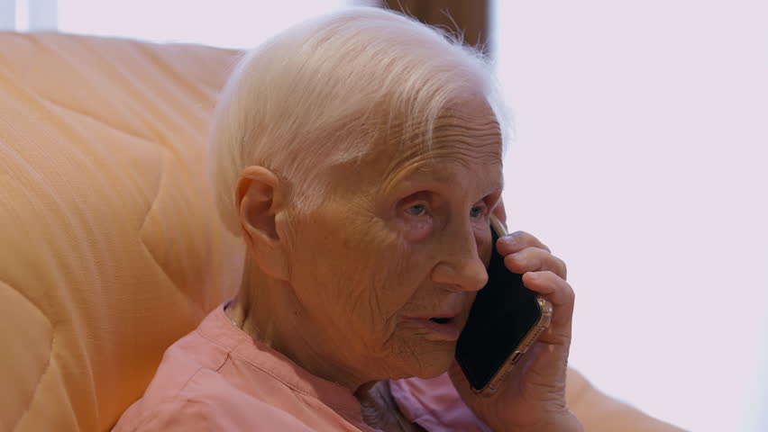 Close-up old Caucasian woman talking on phone sitting indoors at home. Serious senior grey-haired retiree chatting using smartphone. Lifestyle and modern technologies