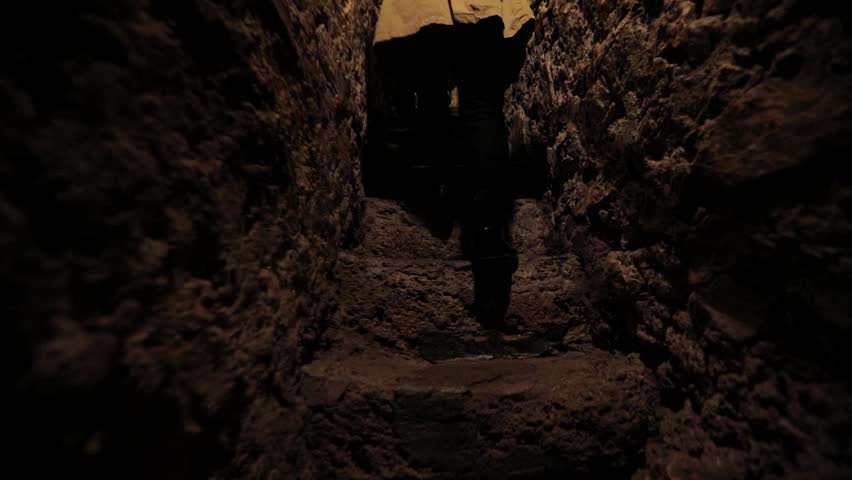 a girl in a raincoat climbs the stairs along a narrow corridor in the fortress. steps of a girl on the catacomb stairs. tunnel in the castle. general plan