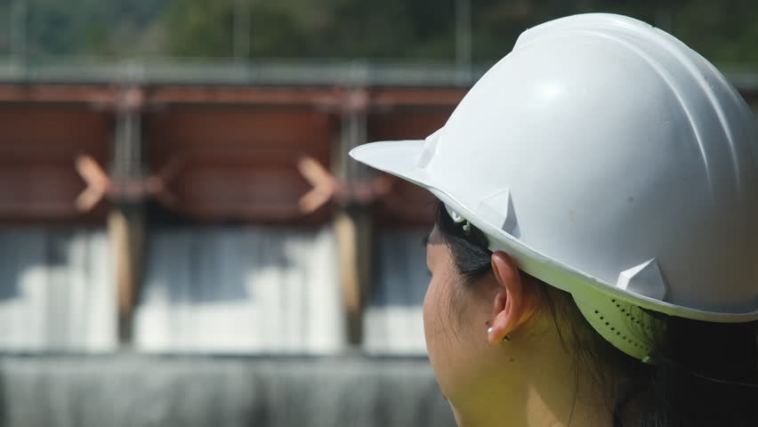 Female engineer wearing green vest and white helmet working outside dam with hydroelectric power plant and irrigation. Renewable energy system. Sustainable energy concept.