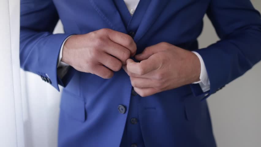 A closeup shot of a bridegroom holding his blue costume while standing near a window | Shutterstock HD Video #1099136205