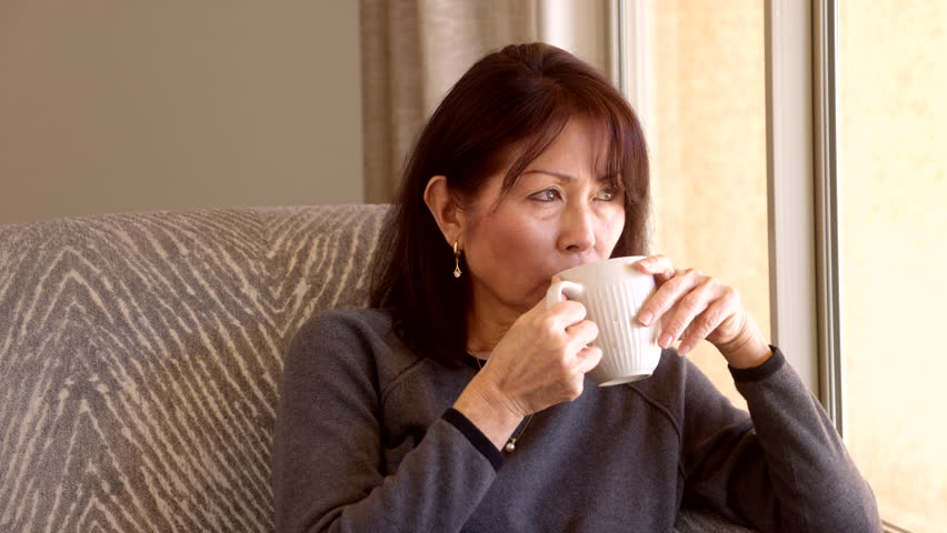 Attractive stylish woman with dimples, brown top and long, dark hair relaxing in comfortable stuffed chair looking out bright living room window drinking coffee. Mature asian is casual and authentic.