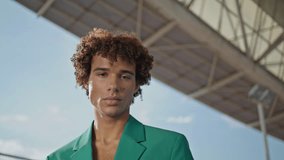 Fashion model posing alone outdoors portrait. Serious young man looking camera at summer street closeup. Curly guy in elegant suit standing stadium zoom out. Casual teenager face in glamour outfit - Powered by Shutterstock - Get 15% off with code: PIKWIZARD15