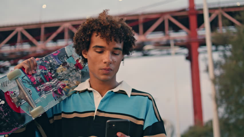 Contemporary skateboarder texting phone outdoors closeup. Curly young man carrying longboard at embankment portrait. Positive guy chatting smartphone on sunset bridge place. Handsome teen walking city Royalty-Free Stock Footage #1099143081