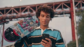 Contemporary skateboarder texting phone outdoors closeup. Curly young man carrying longboard at embankment portrait. Positive guy chatting smartphone on sunset bridge place. Handsome teen walking city - Powered by Shutterstock - Get 15% off with code: PIKWIZARD15