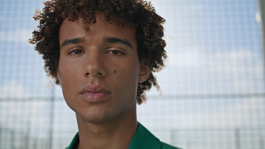 Curly model winking camera at stadium background portrait. Smiling young man posing alone at summer day. Stylish hairstyle guy relaxing closeup. Modern teen face laughing looking directly at street