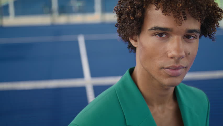 Curly teenager looking lens at stadium background portrait. Serious model posing alone in fashion clothes. Stylish hair guy showing individuality closeup. Young man face relaxing outdoors at summer