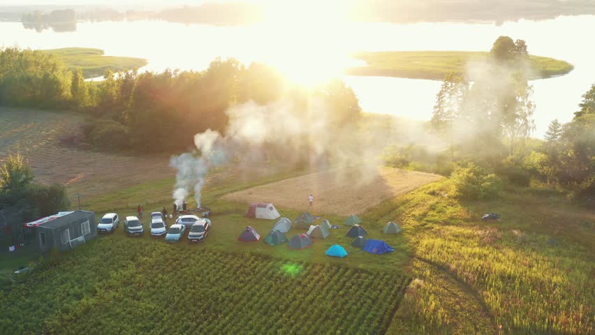 Beautiful aerial shot, camping spot with tents, cars, people and smoke from fire, sunrise over small summer lake shore.