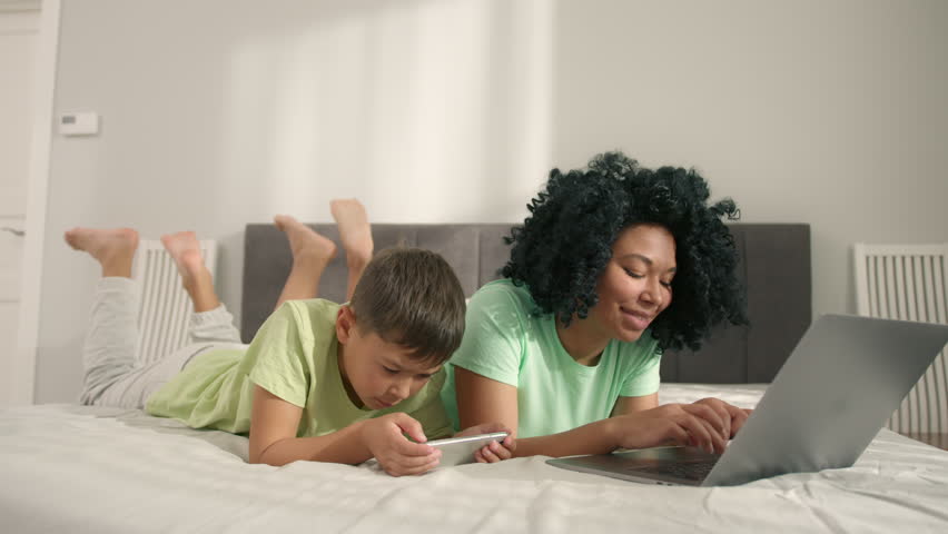 Young generation internet technology addiction.African American woman with her preschool son lying on bed using gadgets. Mom using laptop and boy holding smartphone enjoying online fun video game play Royalty-Free Stock Footage #1099150859