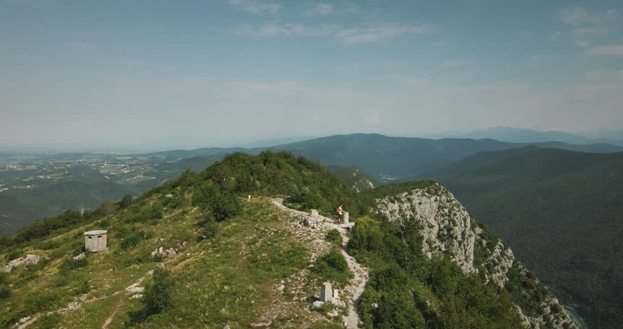 Drone shot of the top of mountain Sabotin where a hiker with an orange backpack stoped by a concrete monument. Great view of nearby mountains, Slovenian side and Italian side.