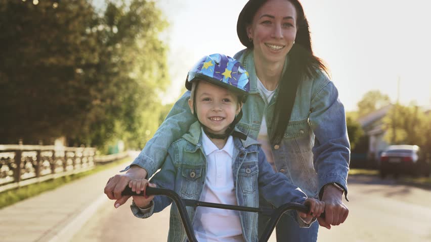 Happy family walk on street the city mother teaches her son child to ride a bike and run nearby. Slow motion woman helps keep balance for safe activity. Sports walk in nature. Healthy lifestyle. | Shutterstock HD Video #1099156111