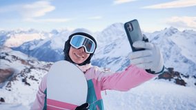 Beautiful woman standing on top mountain in special sports protective clothing: helmet and goggles, taking selfie on mobile phone camera or talking on video call. Winter sport. Snowboard or skis.
