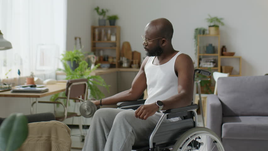 African American man with disability sitting in wheelchair and doing dumbbell curls while exercising at home | Shutterstock HD Video #1099156187