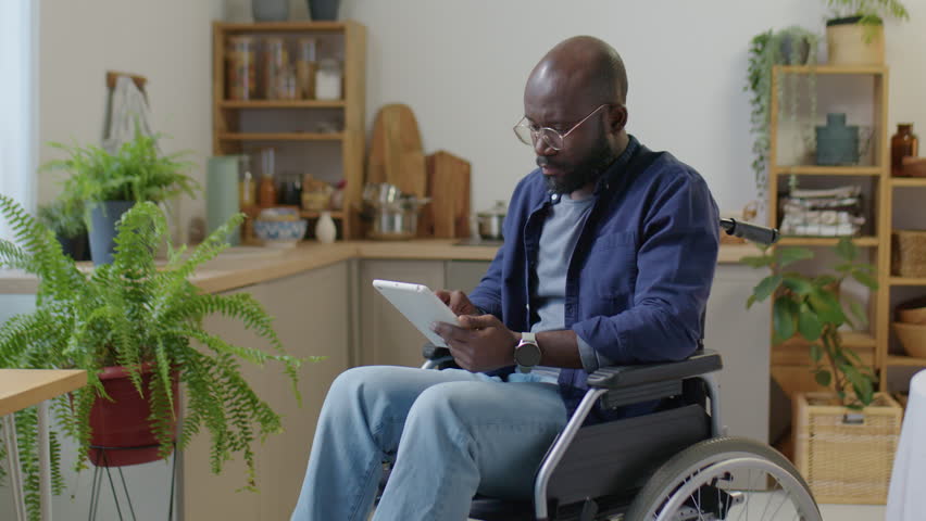 Medium long shot of African American man with disability sitting in wheelchair and using digital tablet at home | Shutterstock HD Video #1099160623