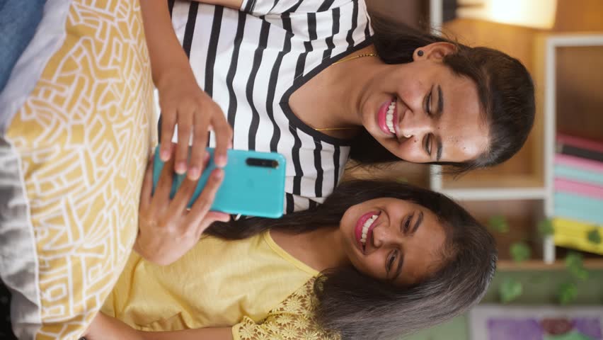 vertical shot of laughing sibling girls while using mobile phone on sofa at home - concept of entertainment, social media and internet connection.