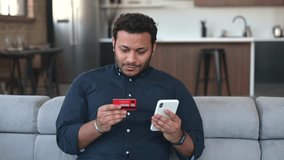 Smiling indian man holds banking card and smartphone, making purchases or ordering online, guy using mobile app for booking or renting, paying online with credit card sitting on the coach