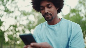 Closeup portrait of young man in light blue t-shirt sitting on city park bench and using his smartphone. Man looks at photos, videos in his mobile phone. lifestyle concept. slow motion