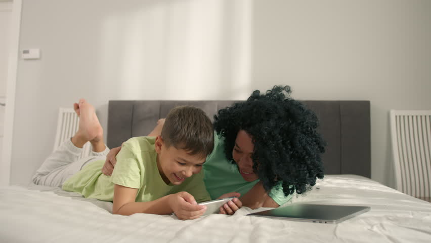 Mother looking how her boy holding smartphone enjoying online fun video game play. Young generation internet technology addiction.African American woman with preschool son lying on bed using gadget Royalty-Free Stock Footage #1099179883