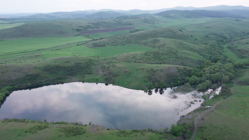 Lake with clear water is located between the hills greened with grass in the Balkan mountains under a cloudy sky