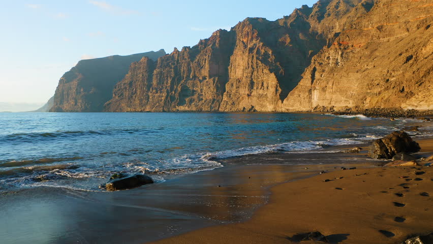Sandy beach with high rocks steep cliffs Los Gigantes Playa de los Guios in Tenerife Island. Tourist attraction. Ocean waves foaming crashing on black volcanic shoreline. Amazing landscape. | Shutterstock HD Video #1099183385