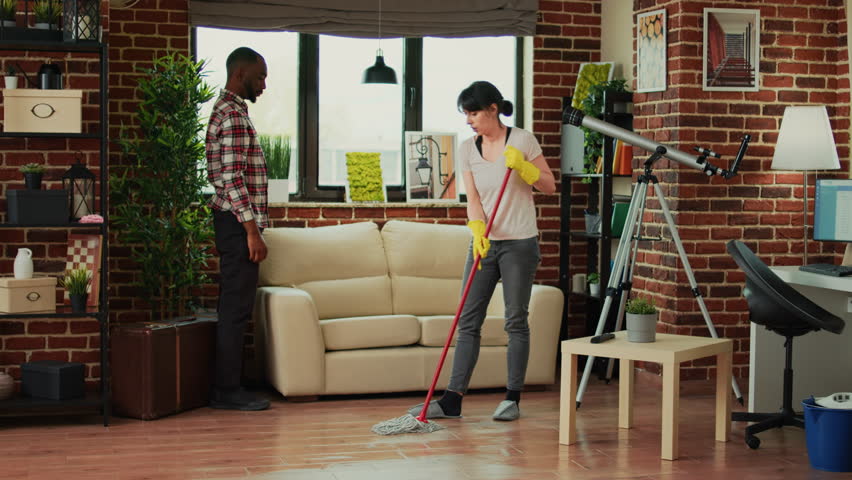 Young couple washing wooden floor with mop, husband helping wife to clean under the couch. Man and woman doing spring cleaning in living room, using all purpose cleaner and washing solution. | Shutterstock HD Video #1099186093