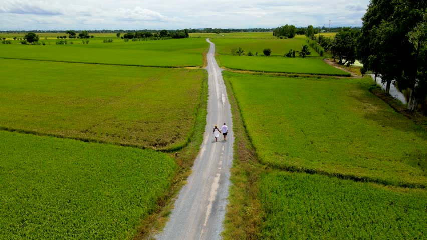 A couple walks on a road with green rice paddy fields in the countryside of Thailand.