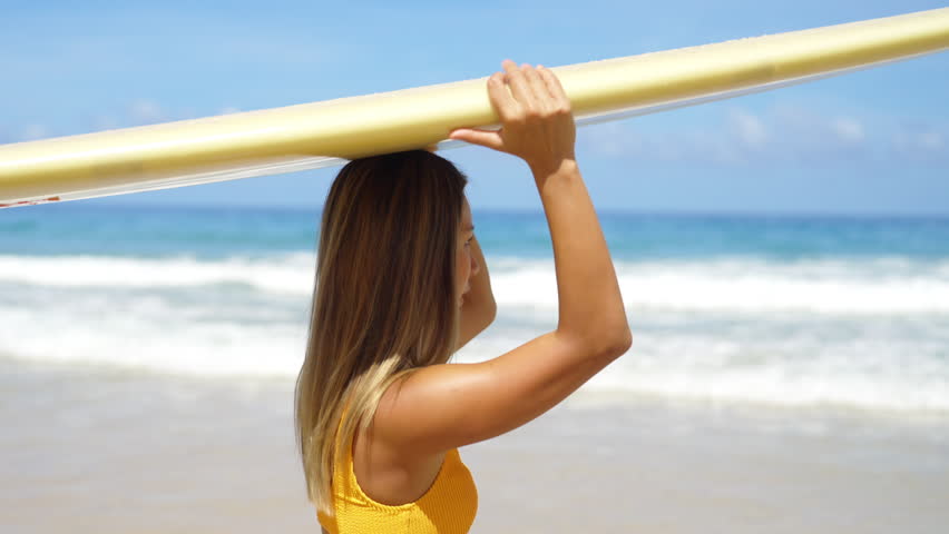 4K Asian woman surfer in bikini holding surfboard walking to the ocean on tropical beach in sunny day. Attractive girl enjoy outdoor activity lifestyle water sport training surfing on summer vacation.