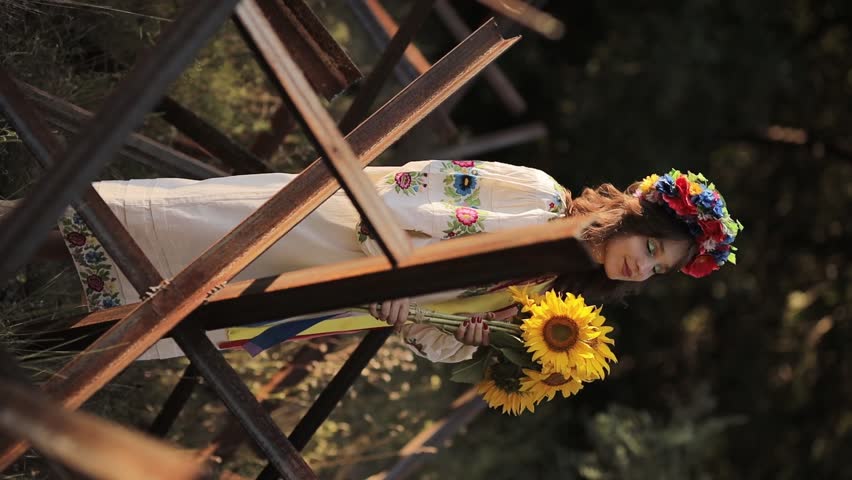 A beautiful Ukrainian girl in a Ukrainian national vyshyvanka dress with a bouquet of sunflowers against the background of anti-tank hedgehogs. war in Ukraine. War of Russia against Ukraine.