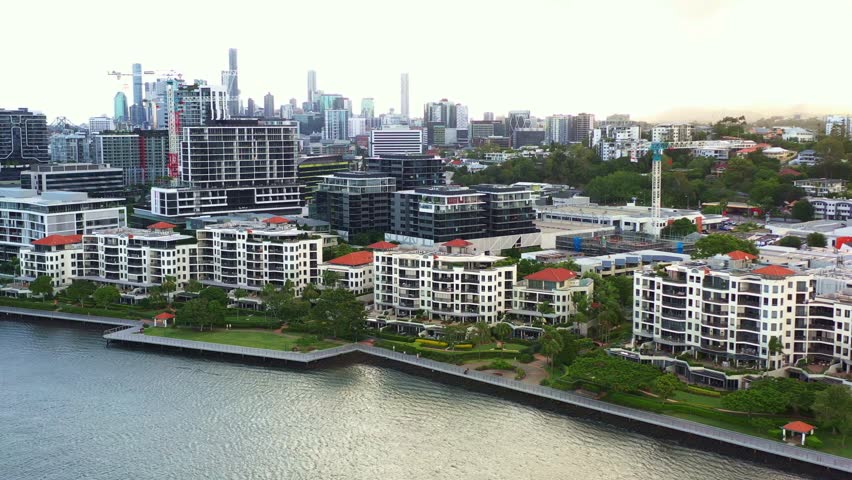 Cinematic aerial view fly around Newstead river terrace, waterfront residential apartment complex along Brisbane river with downtown cityscape on the skyline at sunset, capital city of Queensland.