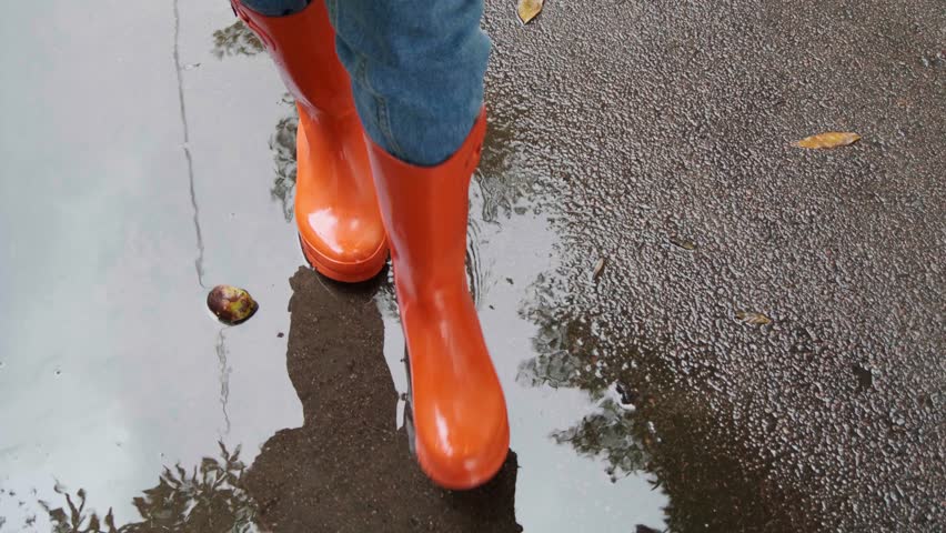 Woman in bright orange rain boots walking through puddles outdoors, above closeup view. Camera moving backwards