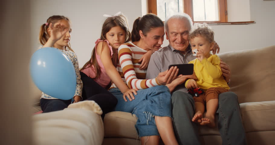 Happy Small Family Having Video Call on Smartphone, waving While Sitting on Sofa in the Living Room. Family of 3 Generations Connecting to a Distant Relative Through Internet and Technology | Shutterstock HD Video #1099208709