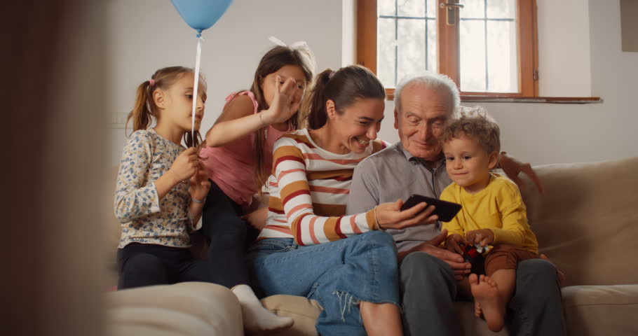 Happy Small Family Having Video Call on Smartphone While Sitting on Sofa in the Living Room. Family of 3 Generations Connecting to a Distant Relative Through Internet, Staying in Touch with Technology | Shutterstock HD Video #1099208711