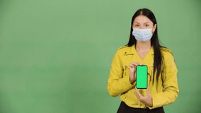 A young beautiful Caucasian woman in a face mask shows a smartphone with green screen to the camera - green screen studio
