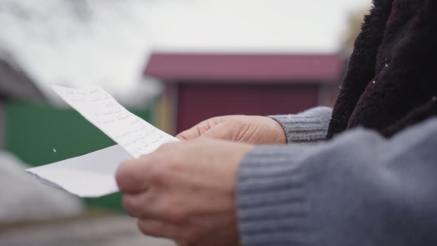 Hands of elderly man holding letter and envelope from friend on blurred background. Pensioner stands in yard on snowy winter day closeup