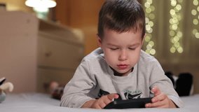 Cute  Little Boy Uses Phone Lying On Bed. Kid Plays Mobile Games And Watch Video On Smartphone