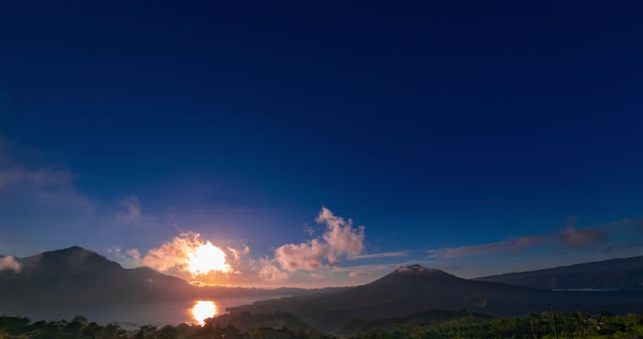 Time lapse of sunrise with view on volcanoes and lake. Clouds are moving quick in blue sky, the sun is rising