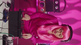 Vertical video: Asian artist with pink hair performing techno music using dj mixer console enjoying to play song with fans, having fun in club at night. Musician doing performance with professional