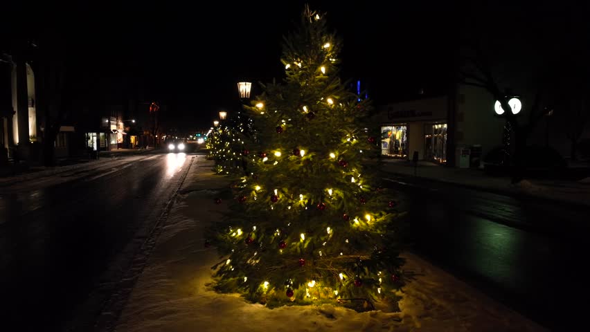 Slow aerial orbit around Christmas tree with lights on main street of Wellsboro Pennsylvania. Car headlights illuminate light snow flurries on cold, dark Christmas morning.
