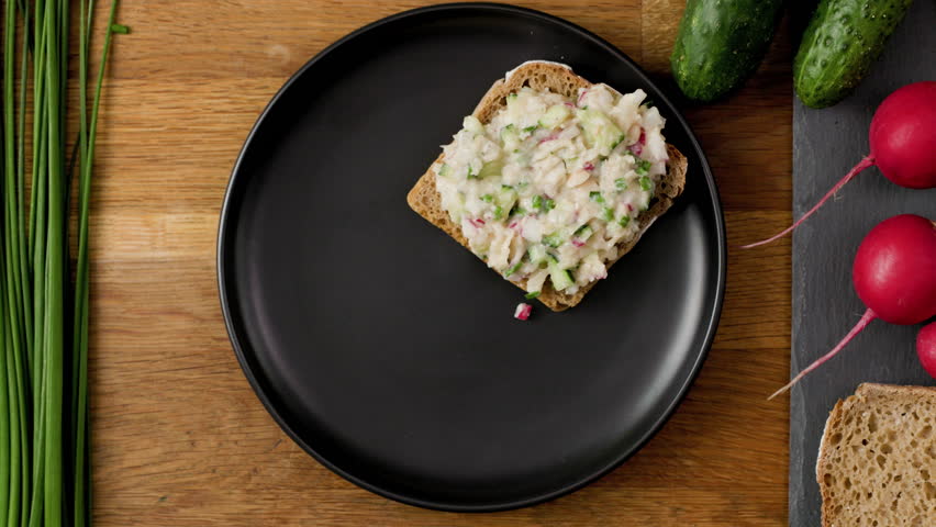 Slather rye bread sandwich with flavorful vegan spread and garnish with slices of radish and chives; dark Scandinavian plate on wooden table; top down, 4k, 25fps | Shutterstock HD Video #1099228703