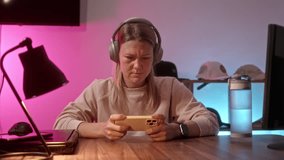 young woman sits at table at home in headphones and play game on phone. neon light in room illuminates place of streamer or avid gamer girl. gamer is passionate about playing on smartphone.