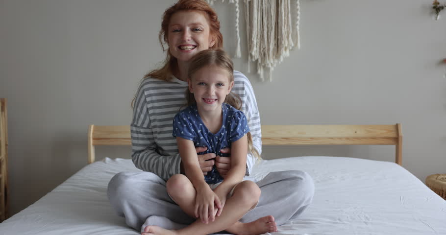 Happy little girl sits on redhead mom laps, smile look at camera. Loving mother tickling adorable cheerful daughter feel overjoyed enjoy playtime sit on bed in cozy bedroom. Family leisure, motherhood Royalty-Free Stock Footage #1099232749
