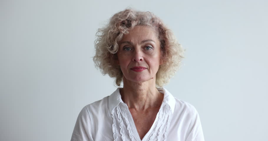 Head shot 55s older blond curly hair female in white casual blouse pose in studio on gray wall background, look at camera having wide toothy candid smile. Generation X or Baby Boomer person portrait Royalty-Free Stock Footage #1099232775
