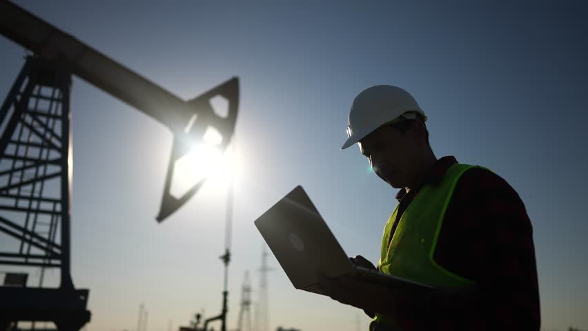 Oil business. a worker works next to an oil pump lifestyle holding a laptop. industry business oil and gas concept. engineer studying the level of oil production on a laptop silhouette at sunset