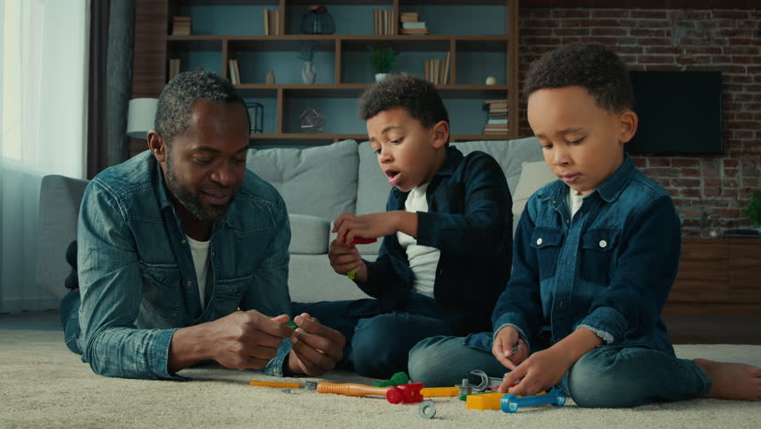 African American ethnic man father with two little preschool sons boys children play game lying on floor at home dad with kids playing with tools toys talking having fun together enjoy funny weekend Royalty-Free Stock Footage #1099249899
