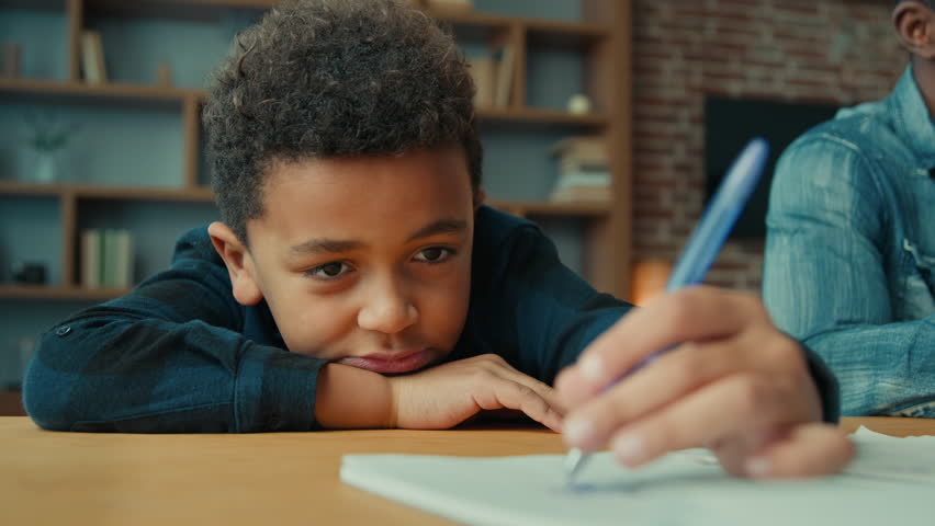 Tired exhausted sad upset fatigued little African American ethnic child boy kid son pupil schoolboy schoolchild lying on table desk writing homework boring class lesson drawing in notebook near father | Shutterstock HD Video #1099249929
