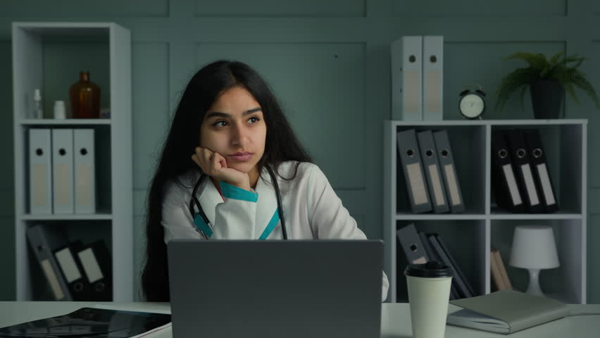 Bored tired woman young practitioner doctor nurse feel sadness lazy female medical worker sit at clinic doing routine work with laptop chatting online use computer app check electronic test results | Shutterstock HD Video #1099249951