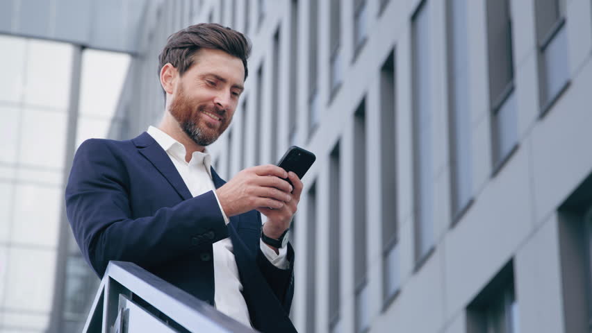 Smiling happy handsome caucasian man holding mobile phone checking email reading message receiving good news successful businessman browsing social network makes order in online store using cellphone Royalty-Free Stock Footage #1099249953
